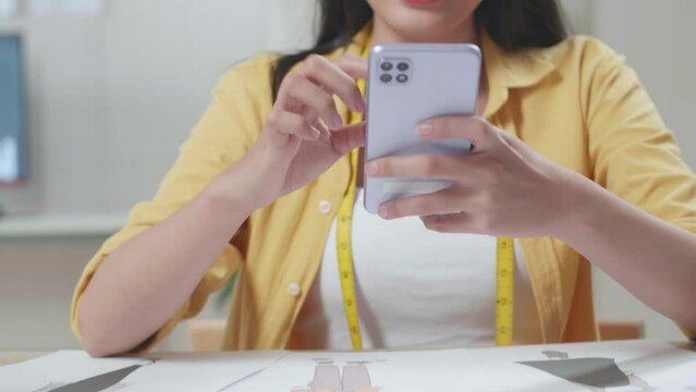 Close Up Of Smiling Asian Female Designer Comparing The Clothes Drawing Pictures On The Table To The Pictures On Smartphone While Designing Clothes In The Studio

