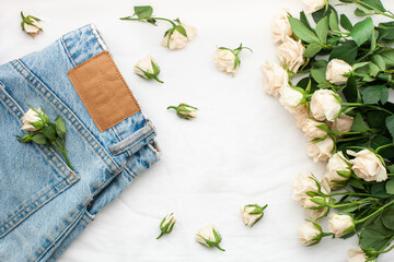 Feminine stylish composition with blue jeans and tea roses on a white bed sheet background. Flat lay, top view. Space for your product or text in the centre (cosmetics, perfume, accessories, etc.)
