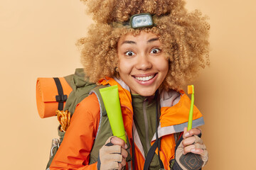 Daily hygiene during camping. Positive cheerful woman with curly hair holds toothpaste and toothbrush smiles broadly spends free time actively enjoys adventure isolated over beige background