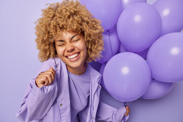 Happy carefree woman with curly blonde hair grins at camera keeps eyes closed dressed in stylish jacket celebrates special occasion holds bunch of inflated helium balloons isolated over purple wall
