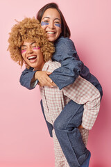 Friendship and fun concept. Joyful diverse female friends give piggyback ride foolish around apply hydrogel patches under eyes undergo beauty procedures dressed in nightwear isolated over pink wall