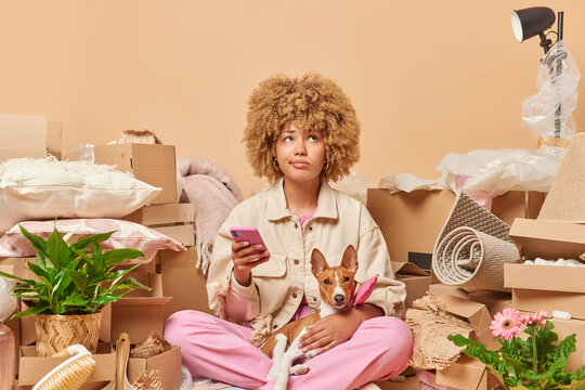 Photo of serious curly haired woman focused above being deep in thoughts holds mobile phone with dog thinks about message content relocates to new house packs things in boxes. Real estate concept