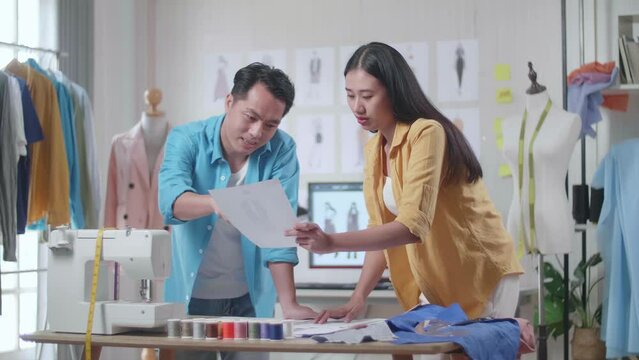 Asian Male And Female Designers With Sewing Machine Discussing About The Clothes Drawing Picture In Hand While Designing Clothes In The Studio

