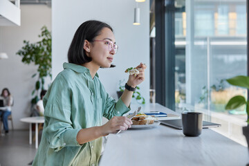 Young ethic woman enjoys eating tasty lunch poses in cozy cafeteria at windowsill surrounded by...