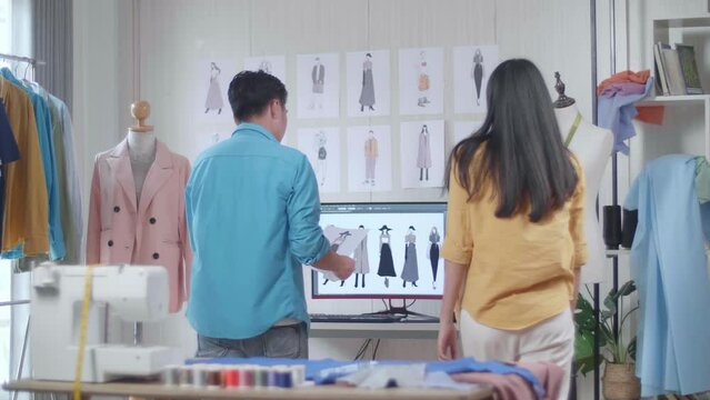 Asian Male And Female Designers With Sewing Machine Walking To Compare Clothes Drawing Picture In Hand To The Pictures On The Wall While Designing Clothes In The Studio
