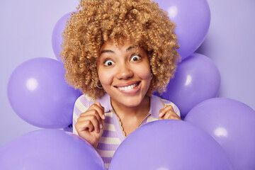 Fototapeta na wymiar Surprised curly haired woman bites lips looks wondered dressed in jumper poses around inflated balloons celebrates special occasion isolated over purple background. People and holiday concept