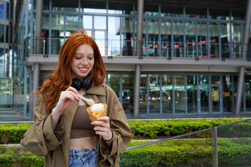 Cool happy teen hipster stylish redhead girl standing in big city urban street eating ice cream. Beautiful teenage generation z girl with red hair wearing trench coat enjoying icecream outdoors. - Powered by Adobe