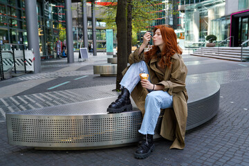 Teen hipster girl eating ice cream sitting on big city urban street. Pretty teen generation z girl with red hair enjoying tasty delicious icecream street dessert spending time outdoors. - Powered by Adobe