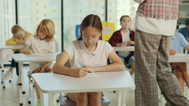 Beautiful brunette asian school girl wearing glasses sits at a first desk in the classroom and fills out tests thinks about answers, and the teacher going through on the background.