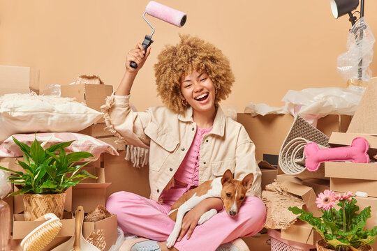 Positive woman holds paint roller does repairment of new house after relocation poses on floor together with pet surrounded by cardboard boxes isolated over beige background. Property and ownership