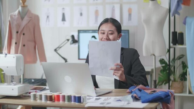 Smiling Asian Female Designer In Business Suit Looking At The Paper In Hand And Comparing It To The Pictures On A Laptop While Designing Clothes In The Studio
