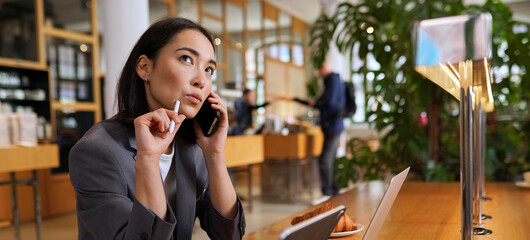 Doubtful Asian young adult business woman talking on cell phone making call working looking away thinking of solution sitting in cafe, considering offer, making decision or feeling doubt.