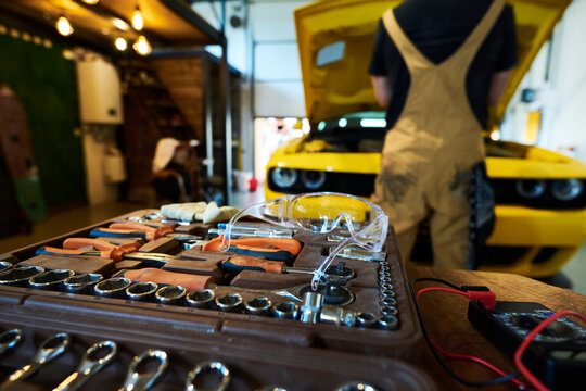 Open toolbox with set of diy handtools and protective eyeglasses on workplace of repairman against mechanic checking car engine