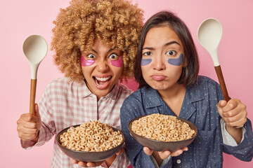 Two diverse young women pose with bowls of cereals and spoons awake early in morning have outraged...