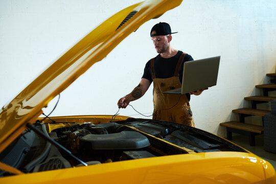 Young mechanic with laptop installing wires into engine of car while carrying out technical diagnostics of motor after repairment