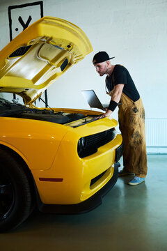 Young repairman or mechanic with laptop standing in front of car with open hood and carrying out technical diagnostics in garage