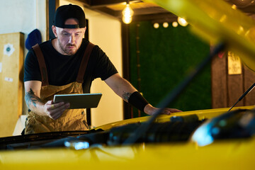 Young worker of maintenance service looking at screen of tablet while bending over engine of car...