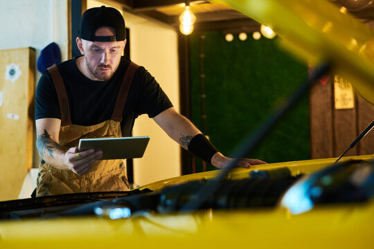Young worker of maintenance service looking at screen of tablet while bending over engine of car and carrying out technical checkup