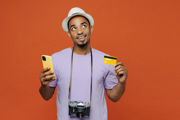 Traveler black man wear purple t-shirt hat use mobile phone credit bank card isolated on plain orange color background Tourist travel abroad in spare time rest getaway Air flight trip journey concept.