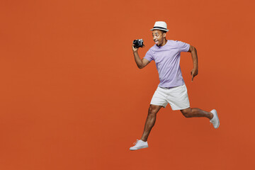 Full body traveler black man wear purple t-shirt hat jump high take photo isolated on plain orange color background. Tourist travel abroad in spare time rest getaway. Air flight trip journey concept.