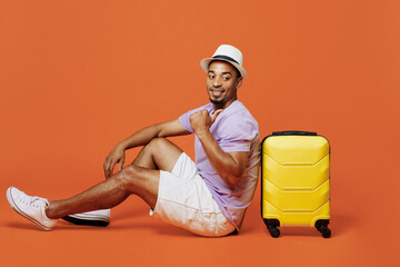 Full body traveler black man wear t-shirt hat sit point finger on suitcase isolated on plain orange color background. Tourist travel abroad in spare time rest getaway. Air flight trip journey concept.