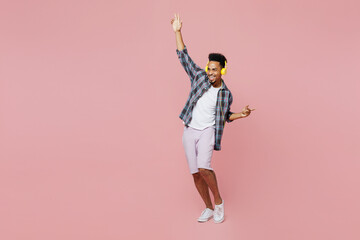 Full size young cool man of African American ethnicity 20s wear blue shirt headphones listen music dance raise up hands isolated on plain pastel light pink background studio People lifestyle concept.