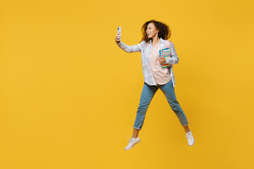 Full size young black teen girl student she wear casual clothes backpack bag hold books jump high use mobile cell phone isolated on plain yellow color background High school university college concept