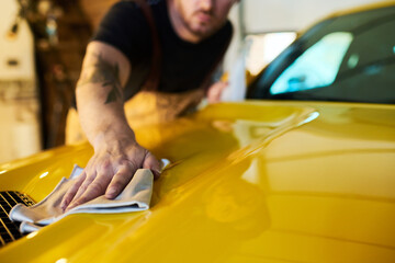 Hand of young male technician with duster wiping surface of yellow car body while cleaning cover of...