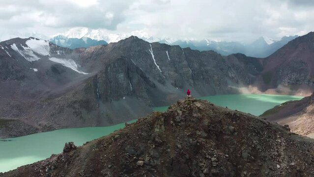 Man in red stay on edge of cliff. Ala-Kul Lake in Issyk Kul Region in Kyrgyzstan. Drone footage 4K. Mountains with big lake and epic clouds. 