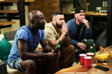 Three young intercultural male friends expressing worry while watching football match broadcast in front of table with beer and snack