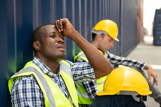African American man worker feel tired after work in container port. Handsome foreman engineer sitting on floor take a break while processes order and product at warehouse logistic in cargo freight.