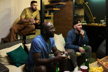Two young men with beer looking at tv screen and watching video game while black guy with joystick...