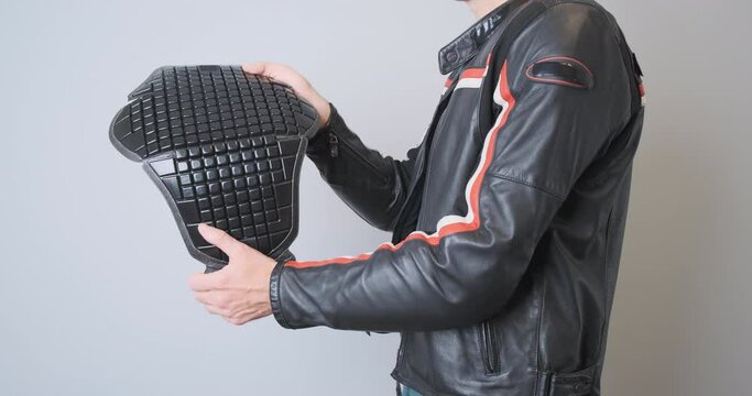 Biker in a leather black sports motorcycle jacket, holds a plastic back protection in his hands, demonstrates it, squeezes it. Side view, close-up, indoors.