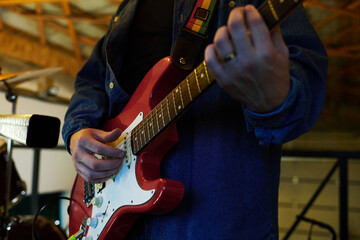 Close-up of young male musician in blue denim jacket plucking strings while playing electric guitar during music repetition in garage