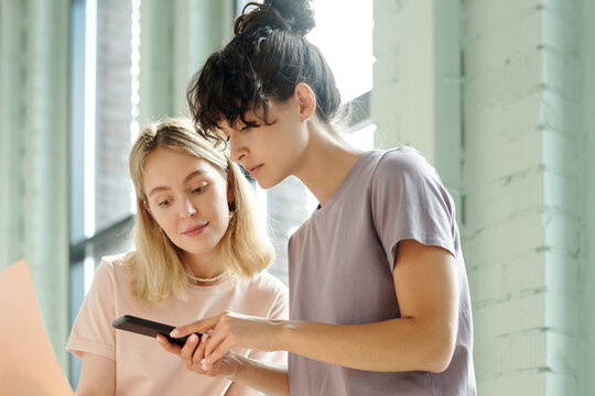 Two young pretty colleagues in t-shirts taking photo of beautycare products on smartphone for advertisement or presentation