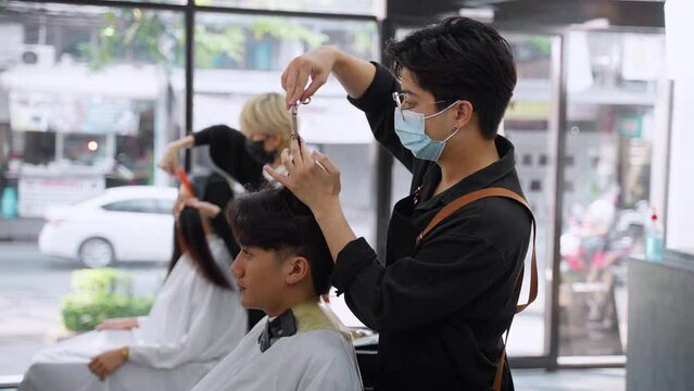 Asian professional male using scissors cutting customer hair in salon. Attractive hairdresser man using scissors cut hair young men and looking at mirror with smiling. Beauty salon business concept.
