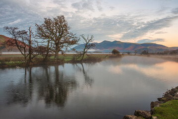 Obraz na płótnie Canvas Absolutely stunning vibrant Autumn sunrise landscape image looking from Manesty Park in Lake Distict towards sunlit Skiddaw Range with mit rolling across Derwentwater surface