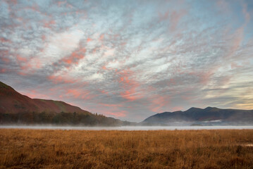 Absolutely stunning vibrant Autumn sunrise landscape image looking from Manesty Park in Lake Distict towards sunlit Skiddaw Range with mit rolling across Derwentwater surface