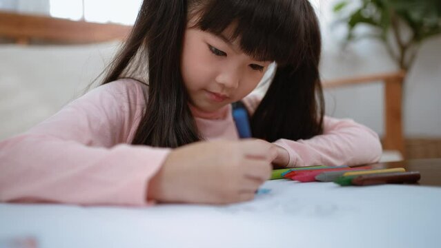 Asian young kid girl coloring and painting on paper in living room. Little adorable child learn how to draw art picture with crayon color paint and brush enjoy creativity activity on holiday at home.