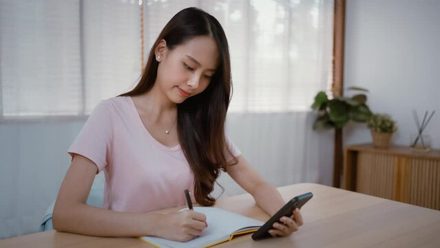Asian young beautiful businesswoman working at home during night time. Attractive freelancer girl sit on study table, use mobile phone call for late work meeting and write note in living room in house