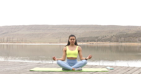 Fototapeta na wymiar American black sportswoman sitting on lake enjoying fresh air meditating for zen, healthcare. Motivation. Active lifestyle.