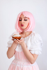 A girl with pink hair and freckles, in a pink dress, holds a festive cupcake in her hands and blows out candles on a white background. Girl makes a wish on her birthday.