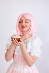 A girl with pink hair and freckles, in a pink dress, holds a festive cake in her hands on a white background. Girl makes a wish on her birthday. Copy space.