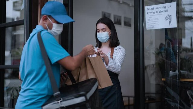 
Asian delivery man wear mask receive food bag from restaurant worker. Attractive business woman waitress giving take a way food bag to delivery food man at coffee shop, New normal lifestyle concept.
