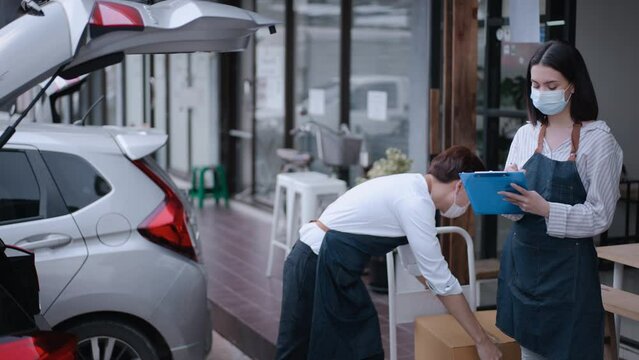 Asian waiter loads parcels into car with waitress check on clipboard. Cafe business owner couple wear mask, work in coffee shop during Covid-19 pandemic . Delivery Purchased transportation concept.