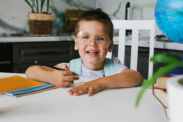 Back to school. One cute toddler boy with glasses is sitting at a desk in home. Kid is learning.