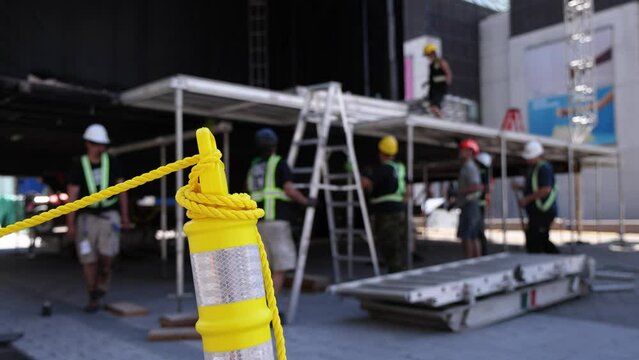 Close up selective focus view of a high visibility bollard on site, as blurry stage builders are seen in background preparing city square for event.