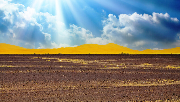 Beautiful wide vast arid dry moroccan desert landscape, black scorched earth, yellow sand dunes, blue dramamtic sky clouds, sun rays backlight - Sahara, Erg Chebbi, Morocco