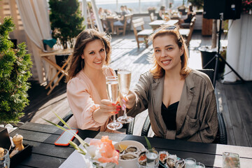 Two Women friends eating fresh oysters and drinking chilled prosecco wine on the summer sunset in...