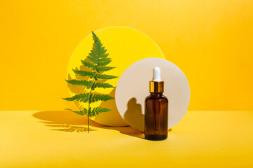 Glass Bottle of serum oil cosmetic product, branch fern on podium pedestal on yellow background. Natural skin care beauty product concept. Mockup for branding